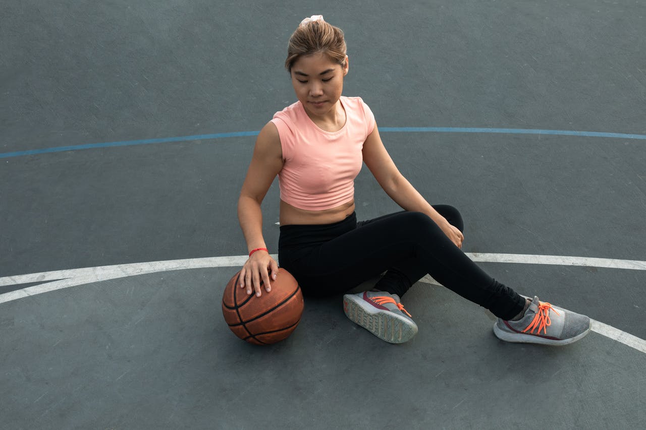 East Asian woman in sportswear sitting on an outdoor basketball court, holding a basketball.